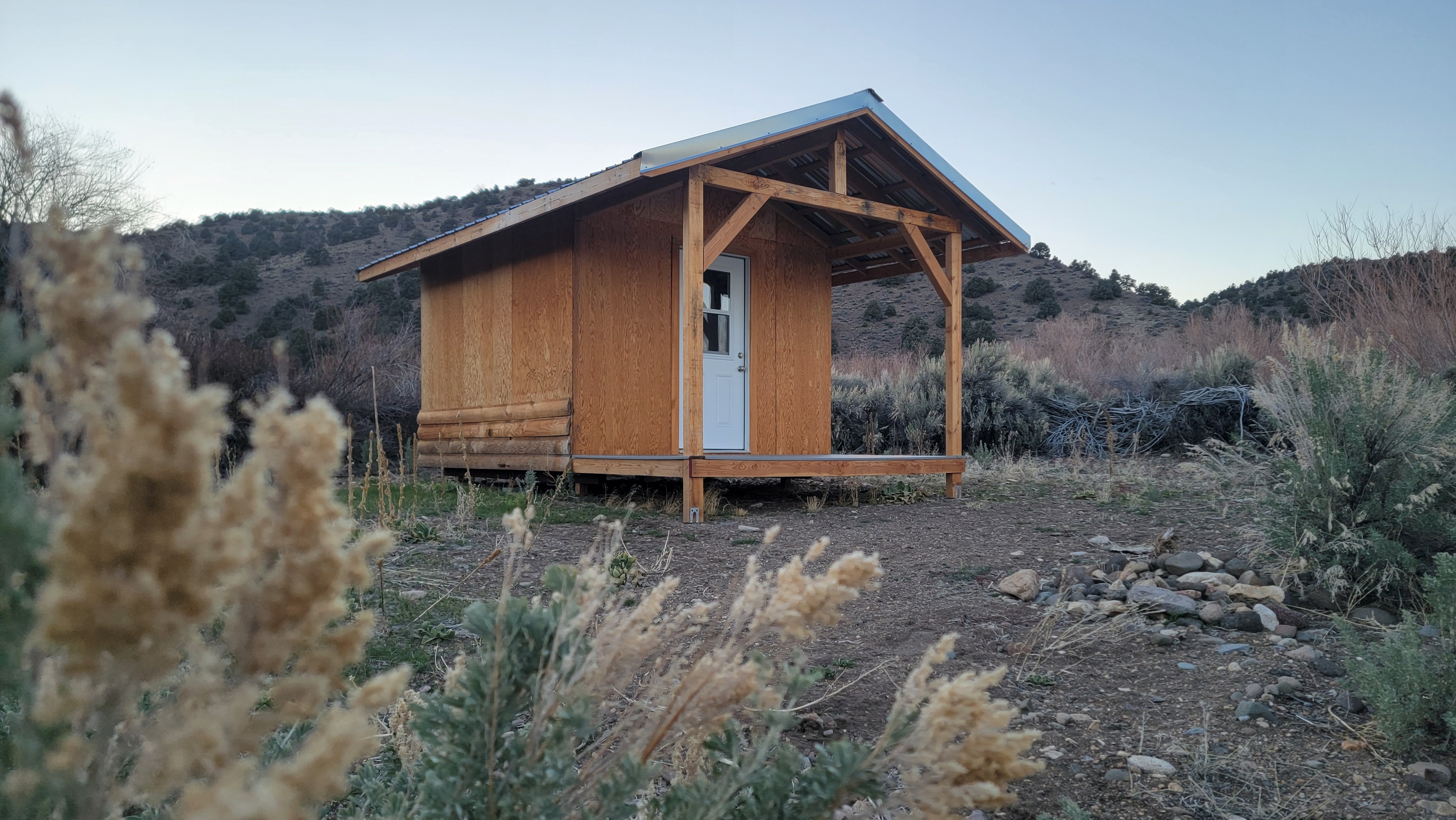 Sweetwater Outpost cabin framed through foreground sagebrush, metal roof, hills behind it