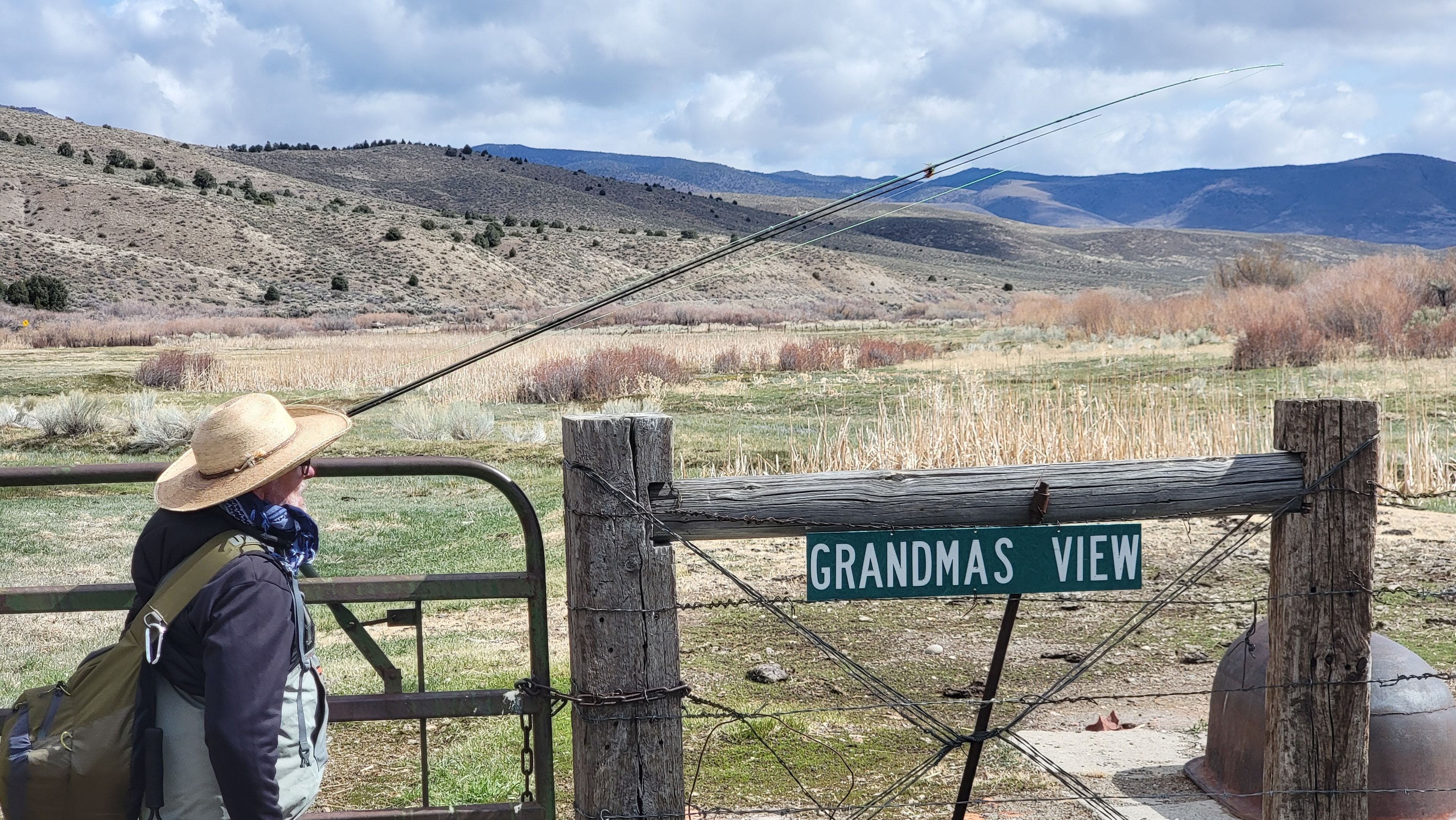 Angler with fly rod arcing overhead leaning on a ranch gate with a Grandmas View sign, East Walker meadow and mountains behind